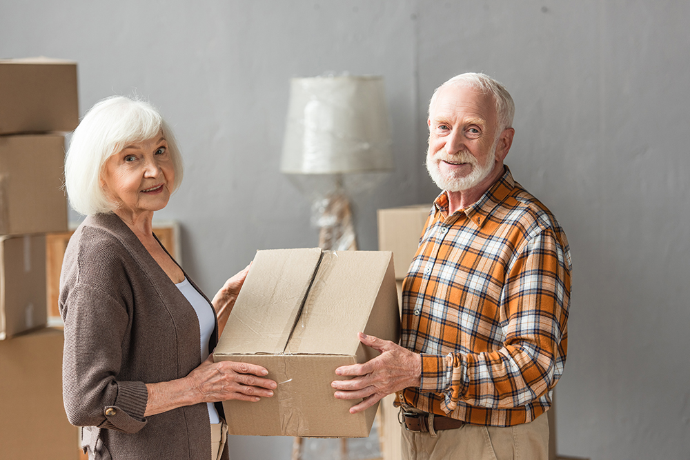senior couple holding cardboard box and looking at camera in new house, moving concept